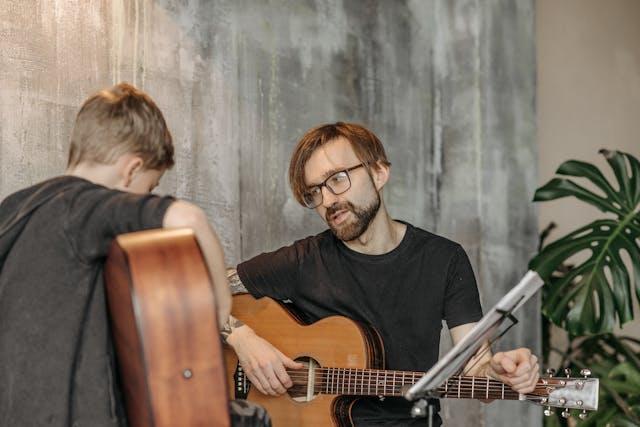 A man with glasses and a beard plays an acoustic guitar while instructing a young boy during a music lesson against a gray textured wall.