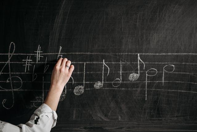 A person’s hand draws musical notes and symbols with white chalk onto a large black chalkboard.